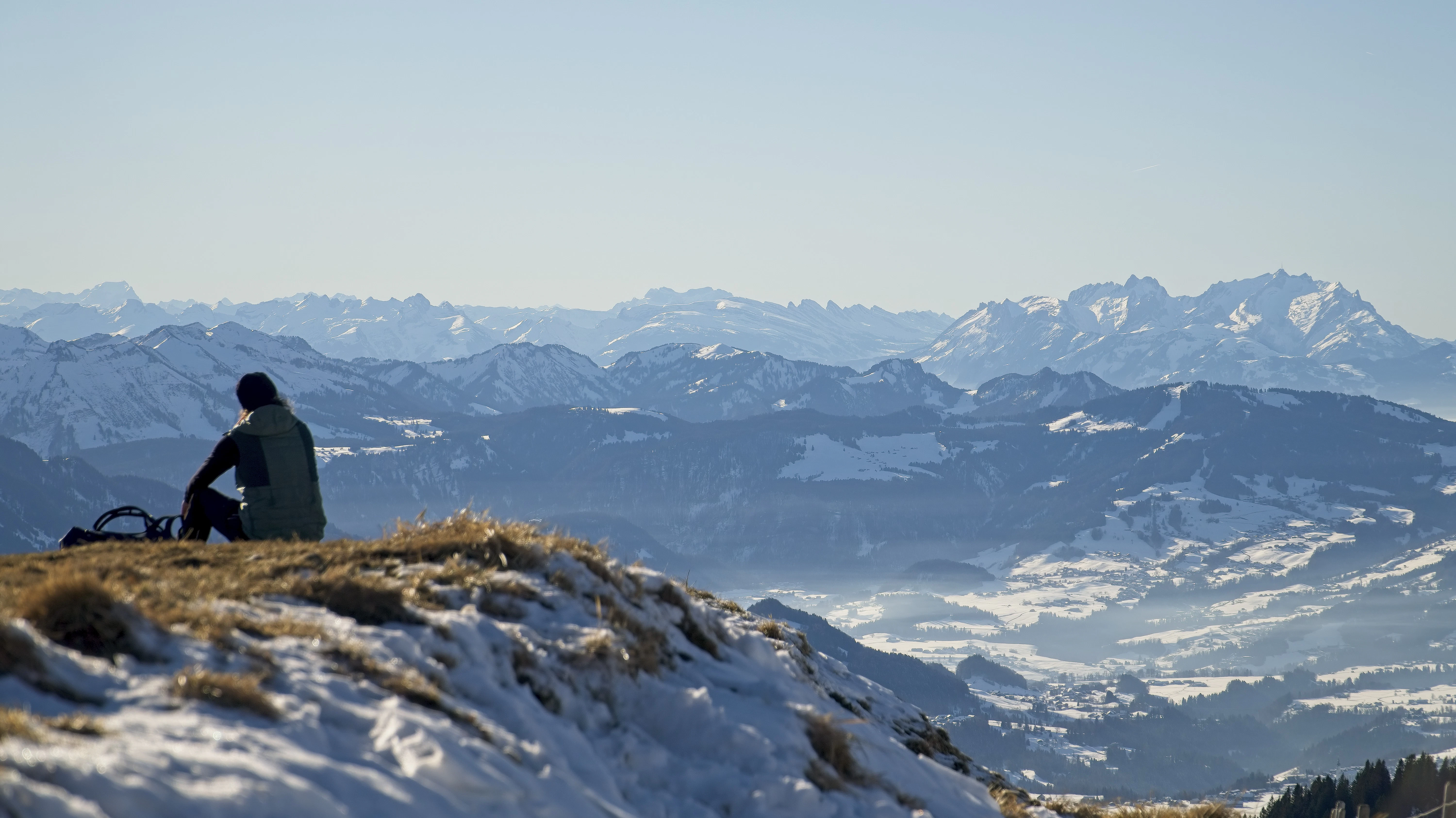 A person sits on a snowy mountain overlooking a valley.
